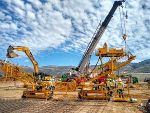 Construction workers, a crane, and other large equipment work under a blue sky and light clouds